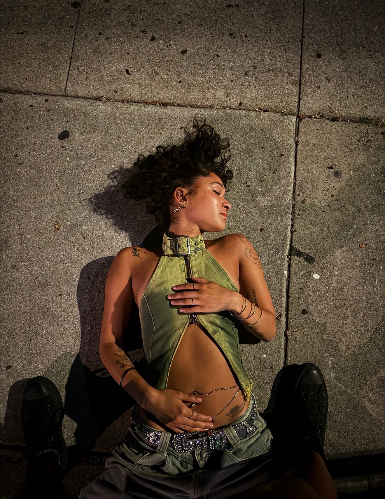 Overhead shot of Maria Williams lying on a night sidewalk in a green denim buckle halter and studded belt, eyes closed