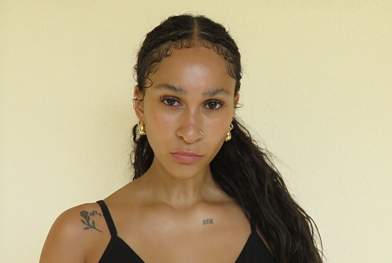 Maria Williams facing the camera against a warm cream wall, wearing a black camisole and gold hoops