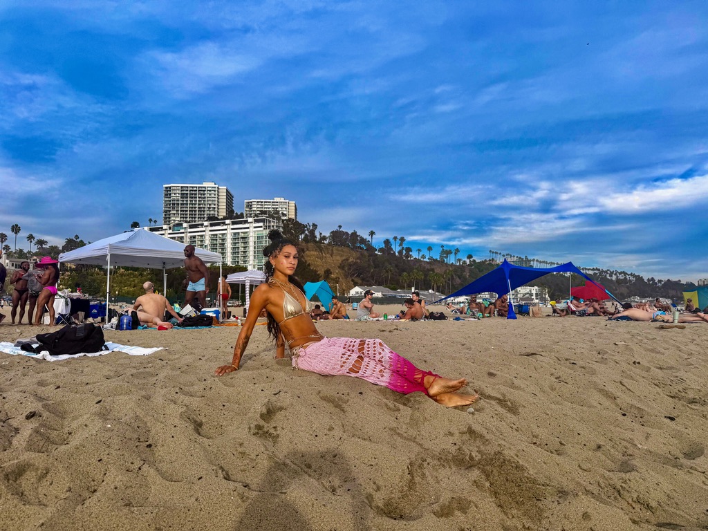 Maria Williams sitting on the sand in a gold bikini and pink crochet skirt on a busy beach