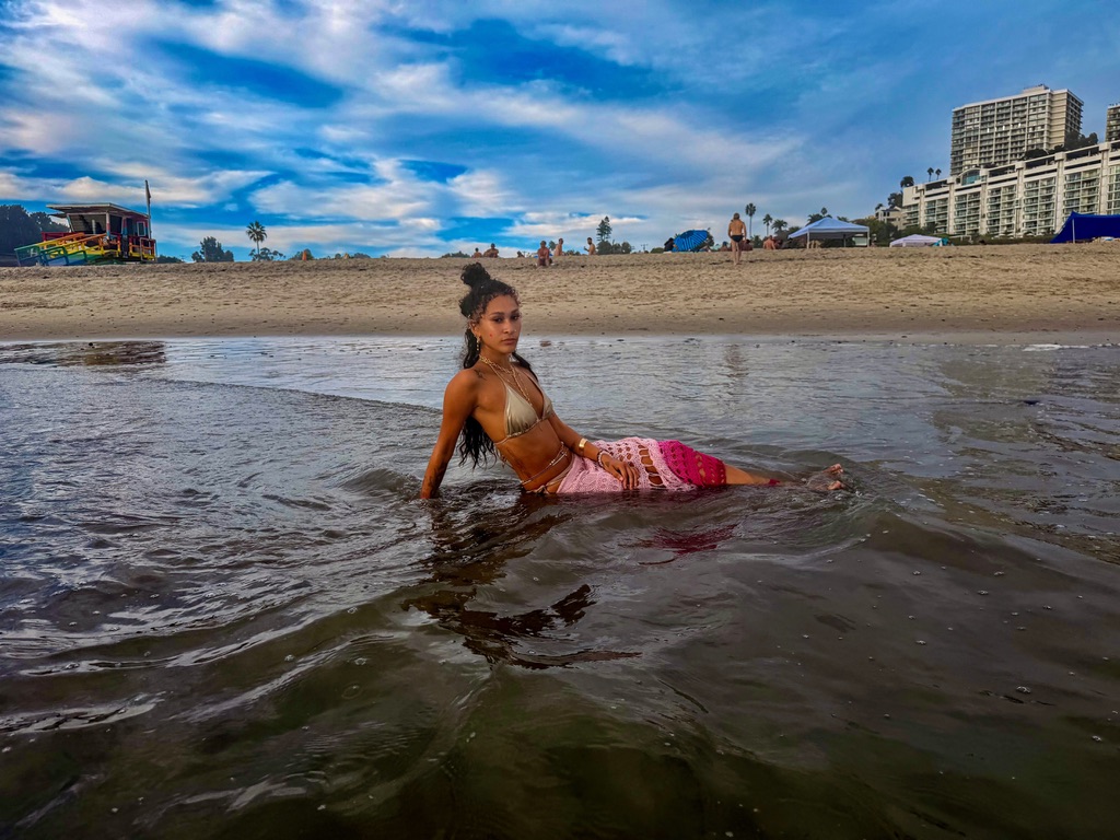 Maria Williams reclining in shallow water in a gold bikini and pink crochet skirt