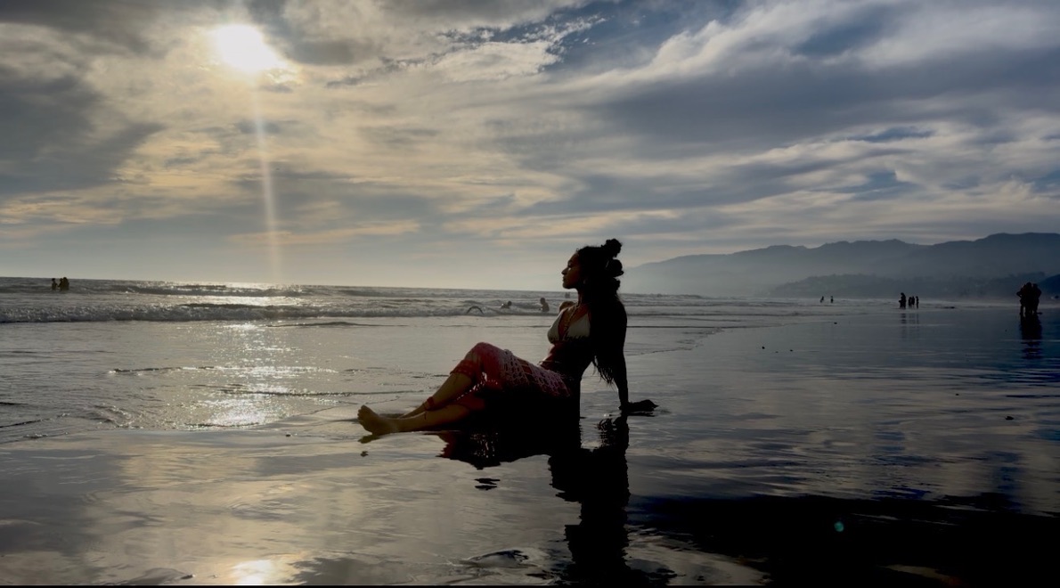 Silhouette of Maria Williams seated on wet sand at sunset with light reflecting off the water