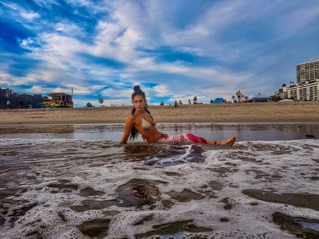 Low angle of Maria Williams lying in the surf as foam rolls in, gold bikini and pink crochet skirt