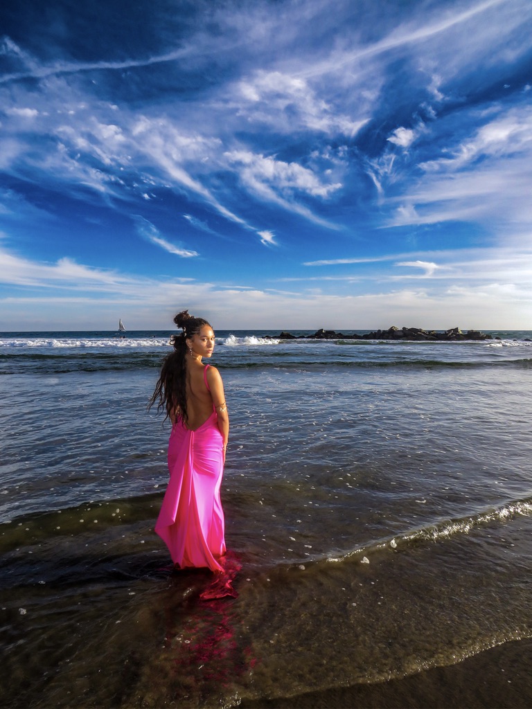 Maria Williams standing in the surf in a pink gown, looking back toward the camera